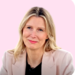 Eine lächelnde Frau mit blonden Haaren und einem hellen Blazer vor einem rosa Hintergrund