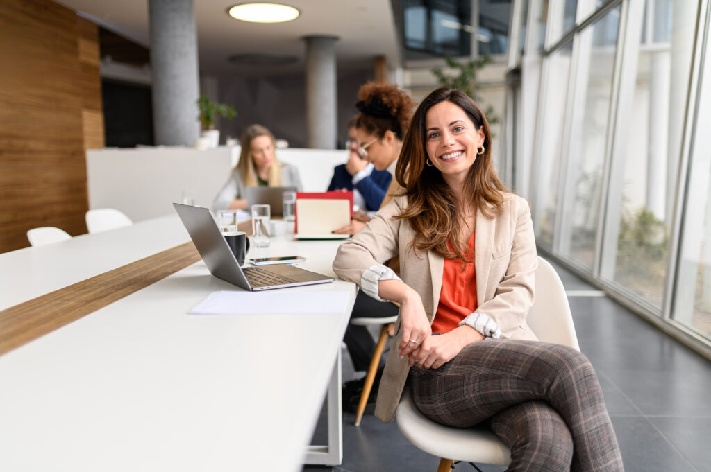 Porträt einer weiblichen Managerin mit Laptop auf dem Tisch, die in einem Besprechungsraum arbeitet, während Kollegen im Hintergrund zusammenarbeiten
