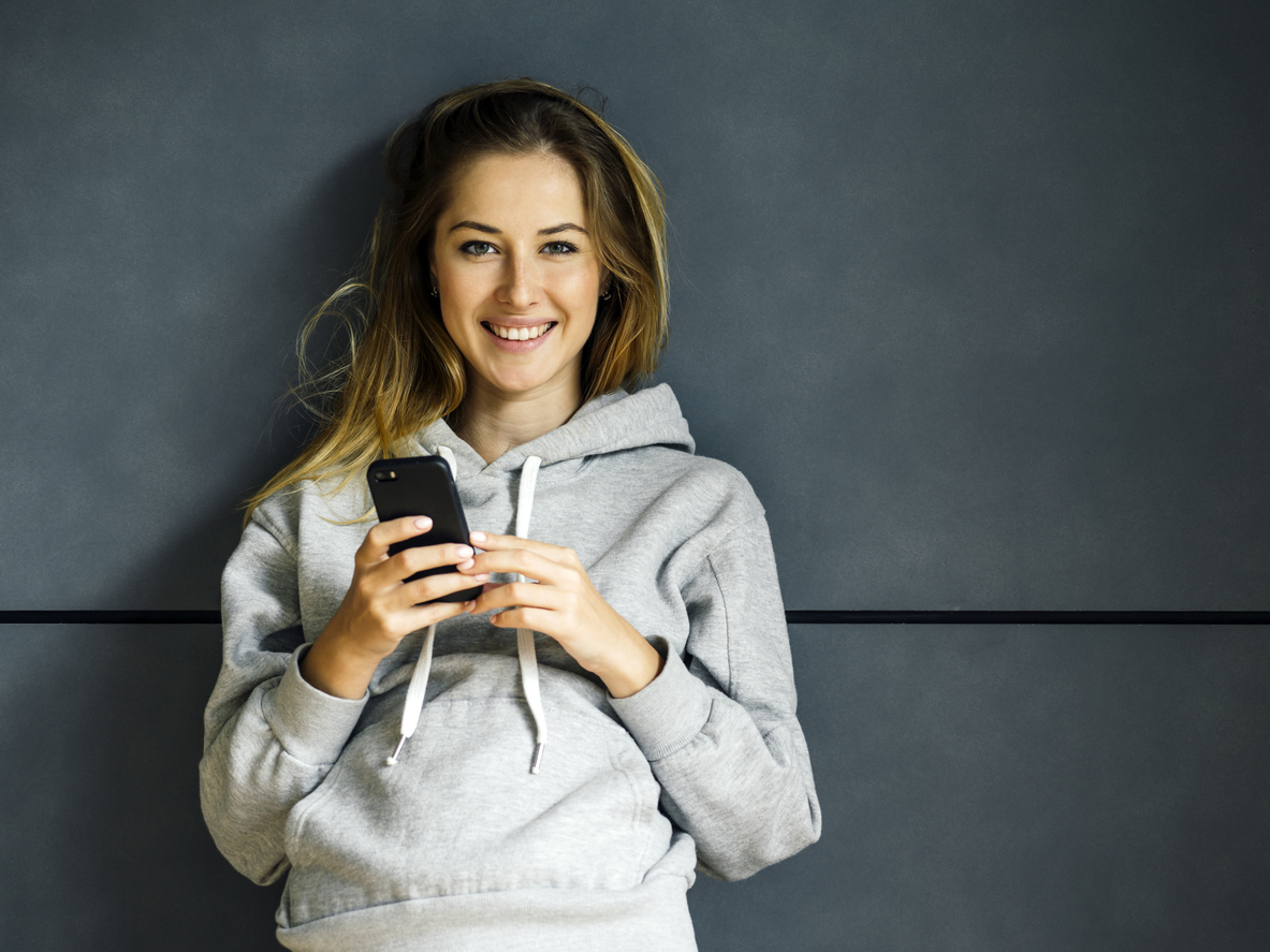 Eine junge blonde Frau, die sich lächelnd an eine Wand lehnt, während sie ihr Mobiltelefon benutzt