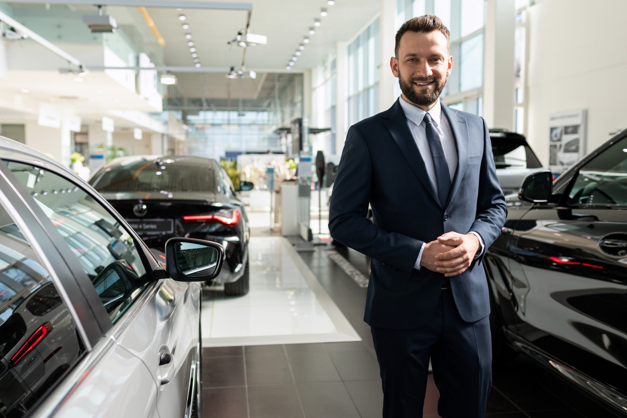 Ein Verkäufer der einen Anzug trägt blickt lächelnd in die Kamera. Im Hintergrund ist der Ausstellungsraum eines Autohauses mit mehreren Fahrzeugen zu sehen.