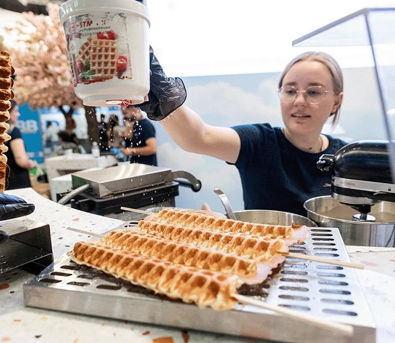 Eine Mitarbeiter*in des Waffelstands bestäubt eine Reihe frisch gebackener Waffeln mit Puderzucker