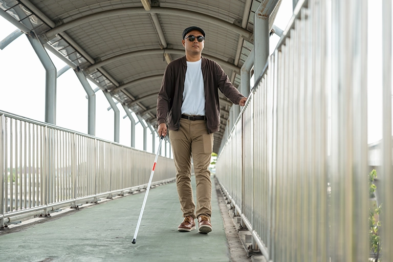 Eine Person mit einem weißen Langstock und einer Sonnenbrille überquert eine Fußgängerbrücke