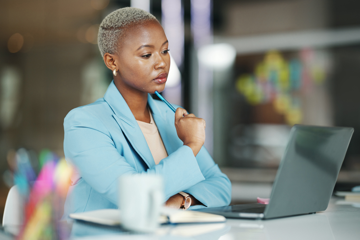 Eine Frau mit einem hellblauen Blazer und kurzen blonden Haaren sitzt an einem Schreibtisch. Sie blickt auf einen Laptop und hat nachdenklich einen Stift an ihr Kinn gelegt.