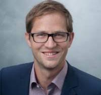 Matthias Schwer hat kurze dunkelblonde Haare und eine dunkle eckige Brille. Er trägt eine blaue Jacke und lächelt in die Kamera